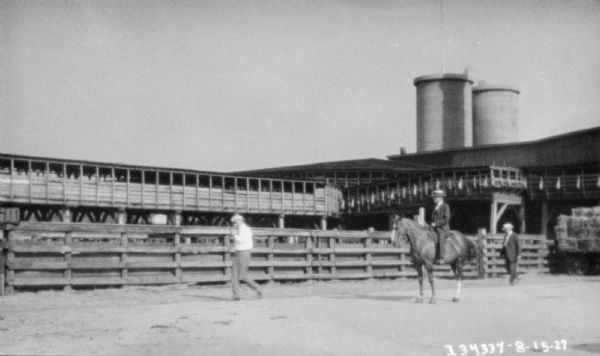 View across grounds towards a man on horseback at a racetrack. In the background are fences, and long, open sided roofed walkways leading to buildings. Smokestacks are behind the buildings.