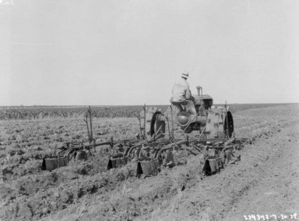 Rear view of a man driving a tractor pulling a cultivator in a field. The man is wearing a jacket that reads: "International" on the back.