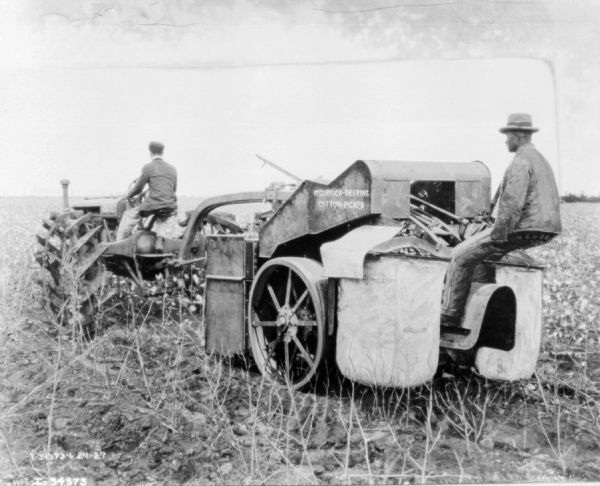 Three-quarter view from left rear of a man driving a tractor pulling another man sitting on the back of a cotton picker.