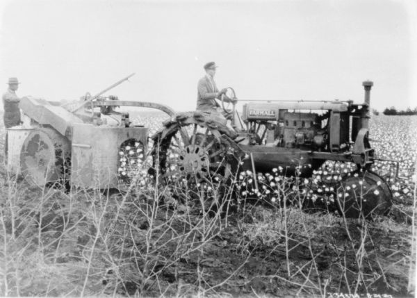 Right side view of a man driving a tractor pulling another man sitting on the back of a cotton picker.