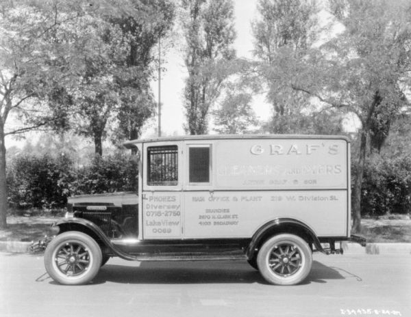 Driver's side view of a delivery truck parked on a street. Behind the truck are bushes and trees along a sidewalk. The sign painted on the side of the truck reads: "Graf's Cleaners and Dyers."