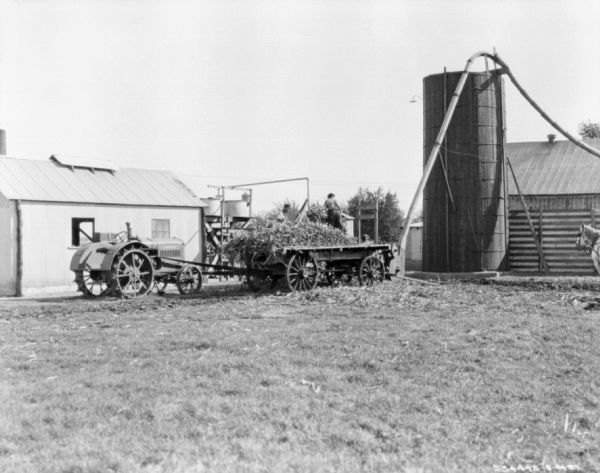 View across barnyard towards men working with a Farmall tractor belt-driving machinery. The men are standing near a wagon piled with cornstalks, and a silo near a barn. There is a horse on the far right.