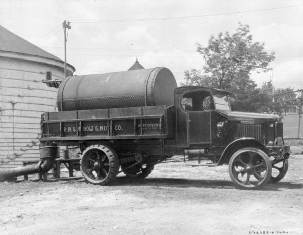 Passenger side view of a delivery truck. The truck has a large storage tank strapped in the truck bed. The sign painted on the side of the truck reads: "R.B. & W? Bolt & Nut Co." There is a round building on the left in the background.