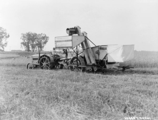 View across field towards a harvester thresher being pulled by a man driving a tractor.
