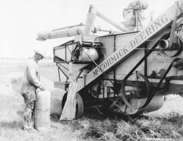 A man is standing in a field next to a threshing machine with a grain sack.