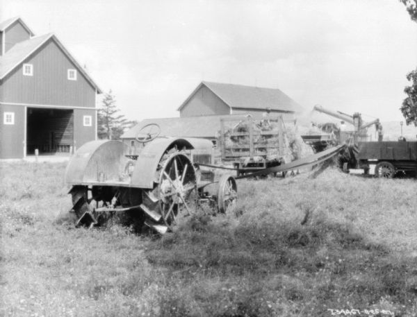 A tractor in the foreground is belt-driving a thresher. A group of men are working near the thresher and wagon. Farm buildings are in the background.