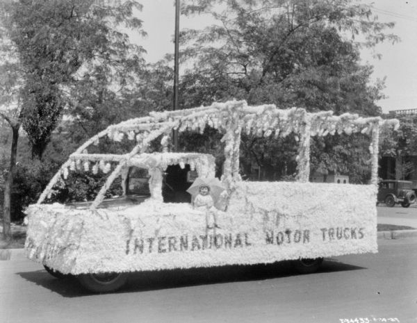 A young girl is sitting on a float holding an umbrella. The decorated float has a sign on the driver's side that reads: "International Motor Trucks."