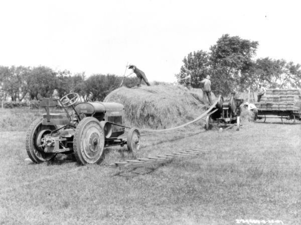 Men working to bale hay. On the left is a tractor belt-driving the hay press, which is set up near men standing on a large hay stack. On the right, two men are working to put the bales of hay on a wagon.