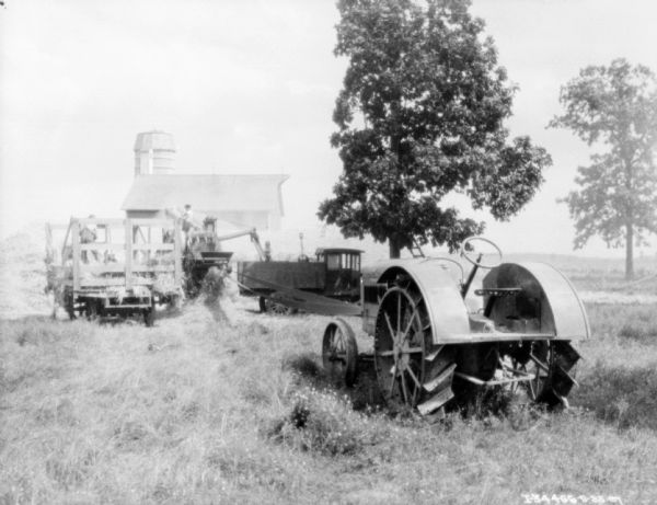 A tractor in the right foreground is belt driving a hay press. There is also what appears to be a thresher, which one person is aiming into the bed of a truck. A wagon piled with hay is on the left. In the background is a barn and silo.