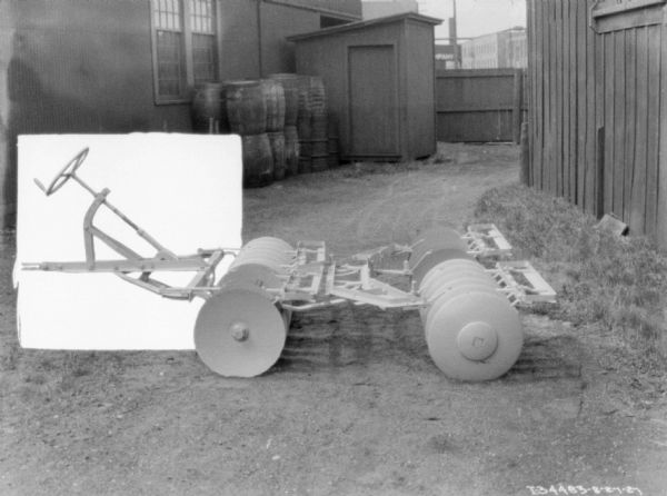 Disk harrow set up outdoors in a yard near buildings. The image has been modified on the left, with a white opaque square behind the steering wheel.