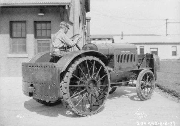 Three-quarter view from right rear of a man driving a McCormick-Deering tractor. Buildings are in the background.