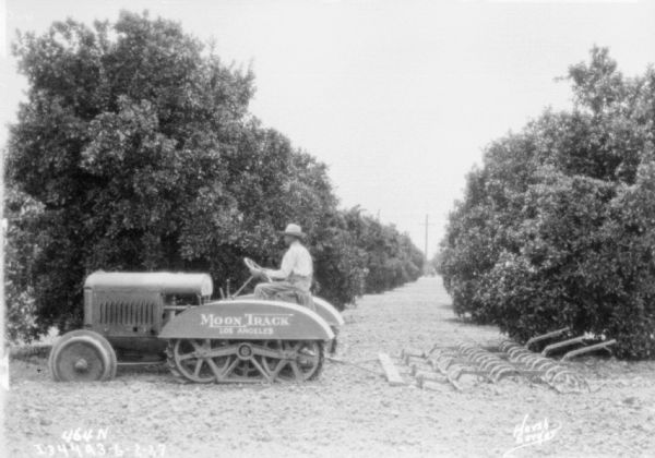 Left side view of a man driving a tractor pulling a spring tooth harrow. Painted sign on the side of the tractor reads: "Moon Track Los Angeles."
