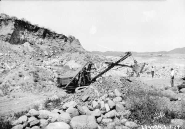 Men working outdoors in a rocky area, perhaps a quarry. One man is operating a steam shovel.