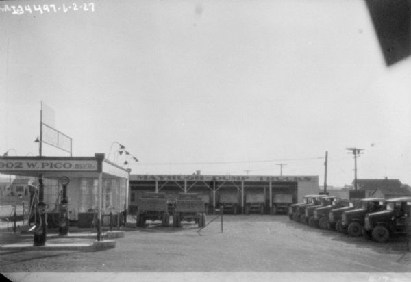 A fleet of dump trucks are parked on a lot and in a building with a sign for "Mayhugh Dump Trucks." There are service pumps near another building on the left.