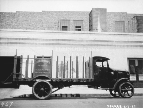 Passenger side view of a delivery truck parked along a curb. Behind the truck is a building with an open garage door. The sign on the side of the truck reads: "Fleischmann's Service Yeast•Diamalt•Arkady•Vinegar."