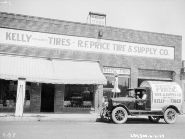 View across street towards a man sitting in the driver's seat of a delivery truck parked along the curb in front of a brick building. Signs on the building and the truck read: "Kelly Tires, Springfield, R.F. Price Tire & Supply."