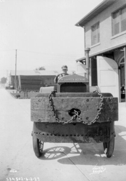 View towards the front of a McCormick-Deering industrial tractor. A man is driving the tractor, and in the background on the right is a two-story building with open garage doors.