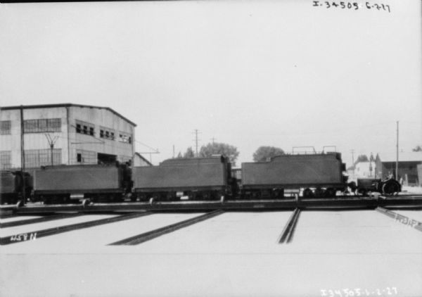 View across railroad yard towards a man driving an industrial tractor on railroad tracks with railroad cars. In the background are industrial buildings.