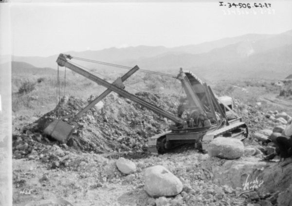 Earthmover working outdoors. A man is standing on the far right. Mountains are in the distance.
