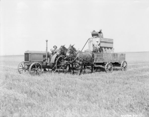 View across field towards a team of two horses pulling a wagon. A man is standing in the wagon. Behind is a man driving a tractor pulling a man is standing on a harvester-thresher.
