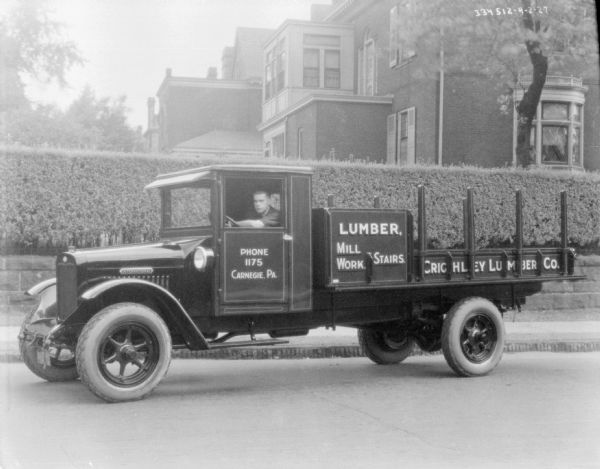 View across street towards a man sitting in the driver's seat of a delivery truck parked along a curb. The sign painted on the truck reads: "Lumbr, Mill Work, Stairs, Crichley Lumber Co." A privet hedge and large buildings are in the background.