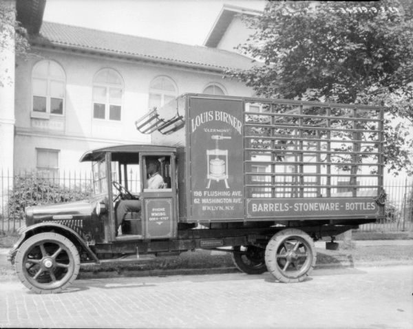 Left side profile view of a man sitting in the driver's seat of a delivery truck. The sign on the side of the truck reads: "Louis Birner 'Clermont' B'klyn, N.Y." and "Barrels-Stoneware-Bottles." There is a large building behind a fence in the background.