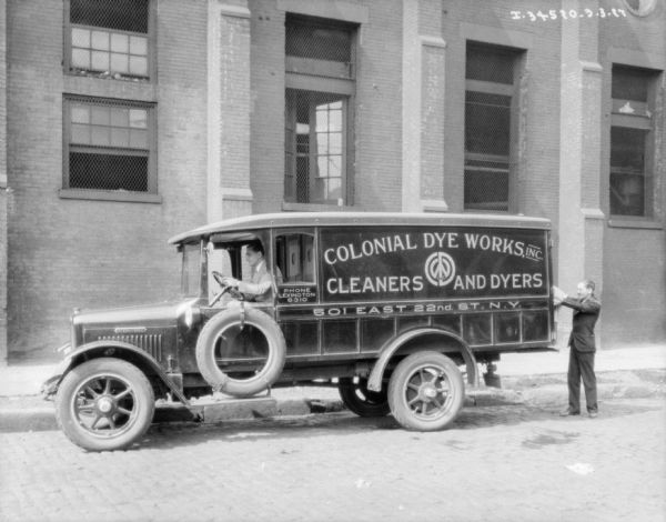 View across street towards a man sitting in the driver's seat of a delivery truck. A man is standing at the back of the truck on the right. The sign painted on the side of the truck reads: "Colonial Dye Works, Inc., Cleaners and Dyers." There is a large brick building behind the truck.
