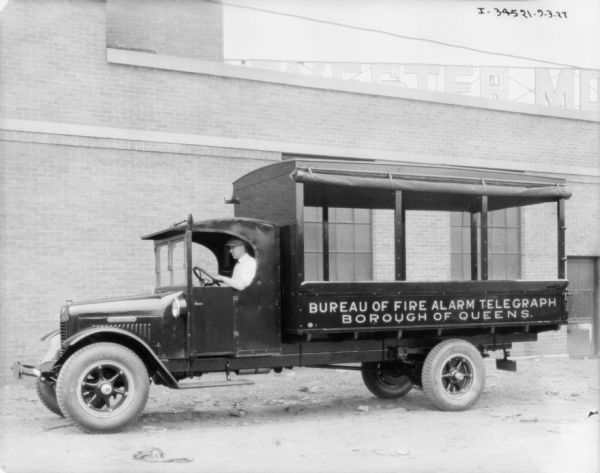 Driver's side view of truck parked in front of a brick building. The canvas on the sides of the truck bed are rolled up. The sign on the side of the trucks reads: "Bureau of Fire Alarm Telegraph, Borough of Queens."