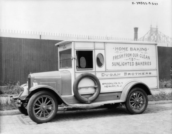 Driver's side view of a bakery delivery truck parked along a curb. The sign painted on the side reads: "Home Baking, Fresh From Our Clean Sunlighted Bakeries, Dugan Bakeries." There is a fence in the background, and beyond the fence is the top of what may be a bridge.