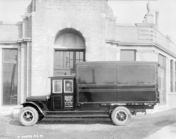 Profile view of driver's side of a delivery truck parked in front of a bridk building, with a sign above the door reading: "International Motor Trucks." The sign on the driver's door reads: "Bigelow Carpet Service Co. Inc."