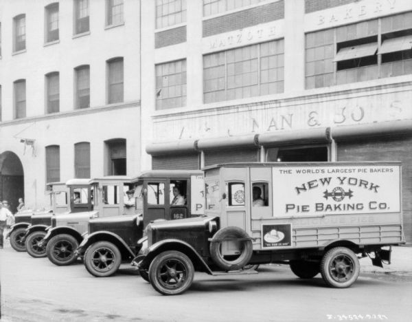 Men are sitting in the driver's seats of a fleet of bakery trucks lined up at an angle in front of a building with a sign on it for Matzoth Bakery. The signs on the side of the trucks reads: "The World's Largest Pie Bakers, New York, Pie Baking Co."
