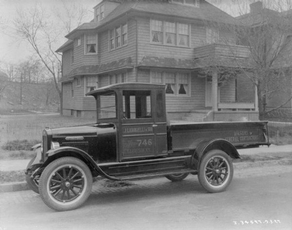 View across street towards a truck parked along the opposite curb. The sign painted on the side of the truck reads: "Masons & General Contractors," and "J. Lambrelli & Son." Houses are along a sidewalk in the background.