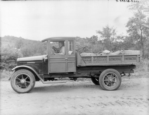 Left side profile view of a man sitting in the driver's seat of a delivery truck parked outdoors. Large rocks are sitting in the open truck bed. The sign on the side of the truck reads: "Henry Corsi, General Contractors."