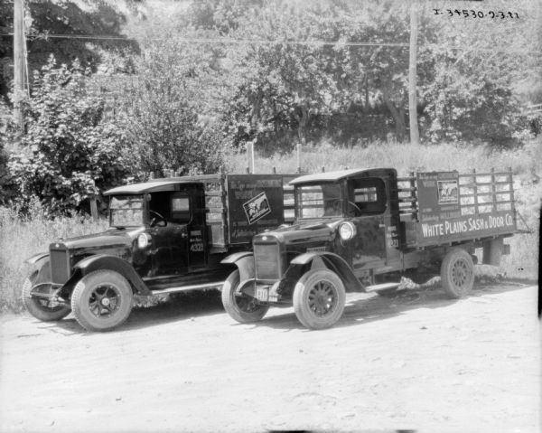 Two delivery trucks with stake bodies are parked together outdoors. The truck on the right has signs painted on the side that read: "White Plains Sash & Door." Another sign on the truck on the left, which is also for White Plains Sash & Door, reads: "This sign on any Construction Work is your guarantee of Satisfactory Millwork."