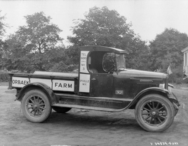 Right side view of a farm truck. There are small American Flags on the front of the truck, and a sign on the back of the truck bed reads: "Orbaek." The corner of a buildings is on the far right, and trees are in the background.