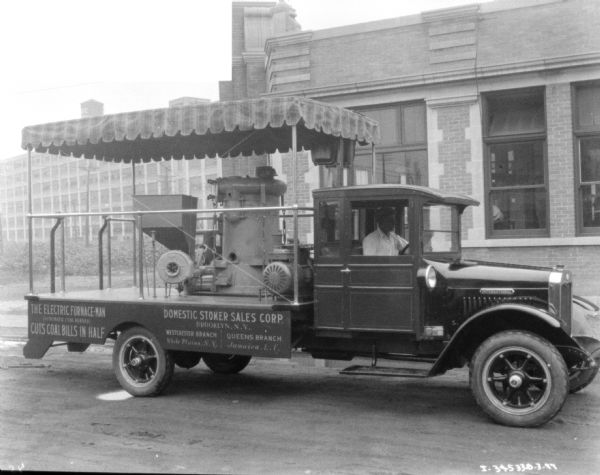 Three-quarter view of right side of truck. A man is sitting in the driver's seat, and the bed of the truck has a striped awning over the top of the truck bed, over an electric furnace. The signs painted on the side of the truck bed read: "The Electric-Furnace-Man (Automatic Coal Burner) Cuts Coal Bills in Half," and "Domestic Stoker Sales Corp, Brooklyn, N.Y."