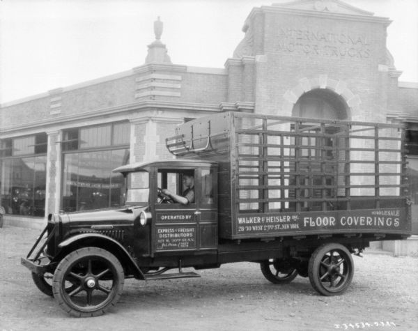 Driver's side view of a truck with a stake body parked in front of a brick building with a sign above the arched entrance that reads: "International Motor Trucks." The sign painted on the side of the trucks reads: "Walk & Heisler Inc. Floor Coverings."
