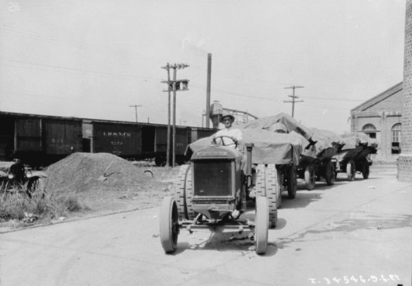 View from front of a man driving a McCormick-Deering tractor to pull three wagons full of supplies. The wagons are covered with tarps. Railroad cars are in the background on the left, and on the right are industrial buildings.