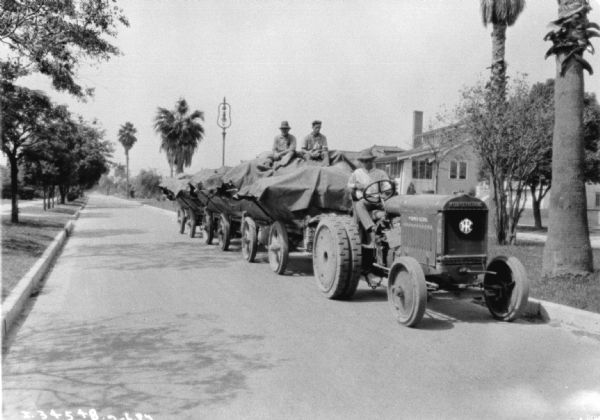 View from front towards a man driving a McCormick-Deering industrial tractor up a street. The tractor is pulling wagons, and two men are sitting on top of the tarps covering the supplies.