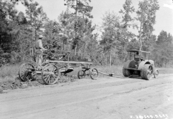 View across unpaved road towards a man driving an industrial tractor pulling another man standing on road construction equipment.