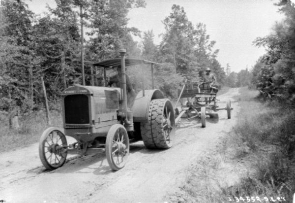 View from side of unpaved road towards the front of an industrial tractor. A man is driving the tractor, and is pulling another man standing on road construction equipment.