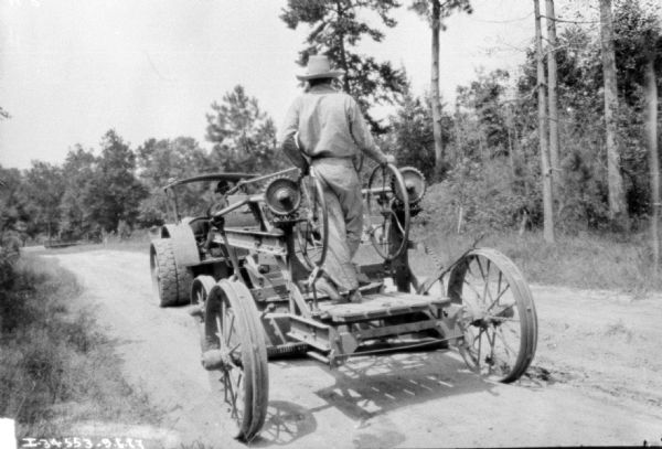Rear view from side of unpaved road towards a man standing on road construction equipment which is being pulled by another man operating an industrial tractor.