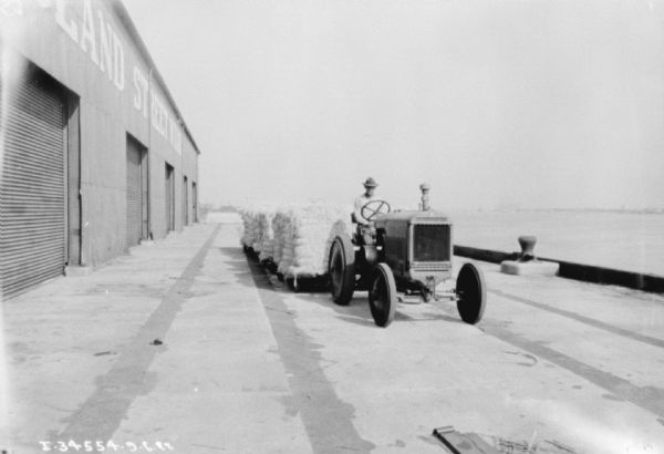 Man driving industrial tractor pulling a long set of carts stacked with bales. The sign on the building on the left reads: "Poland Street Wharf."