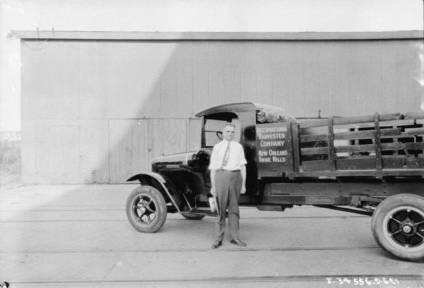 A man is standing next to a truck with a stake body. In the background is an industrial building. The sign on the truck reads: "International Harvester Company, New Orleans Twine Mills."
