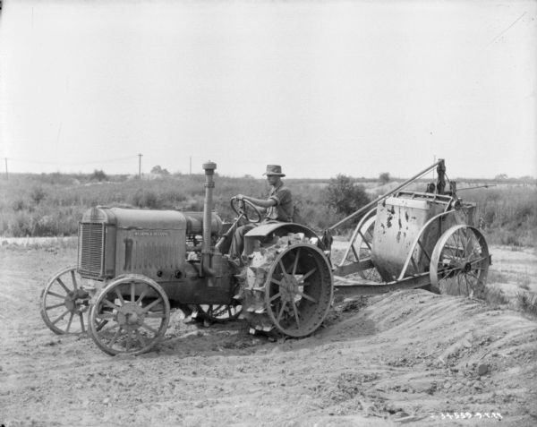 Left side view of a man driving a McCormick-Deering industrial tractor which is pulling an implement on a trailer with large wheels over bare dirt ground.