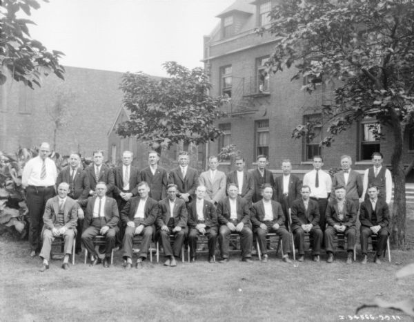Group portrait of men sitting and standing outdoors on a lawn. There are large buildings in the background.