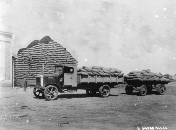 A truck has a truck bed loaded with grain sacks, and is also towing a wagon loaded with grain sacks. There is a large stack of bags of grain under a tarp behind a fence next to a building on the left.