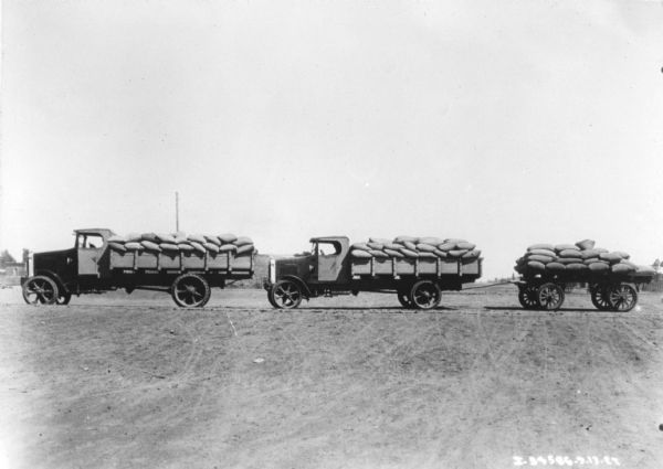 View across bare ground towards men driving delivery trucks loaded with bags of grain. The truck in the center is pulling a wagon also loaded with bags of grain.