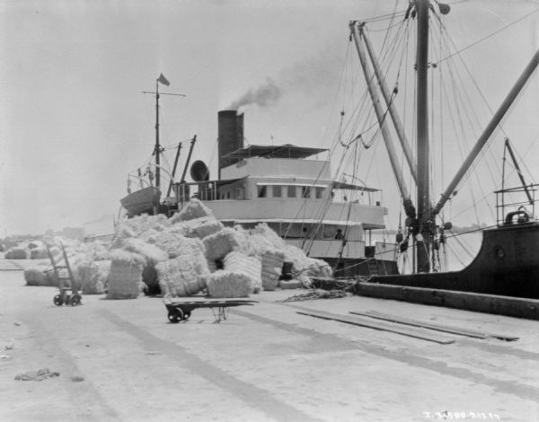 View down wharf towards bales in a large pile. There is a ship at the dock.