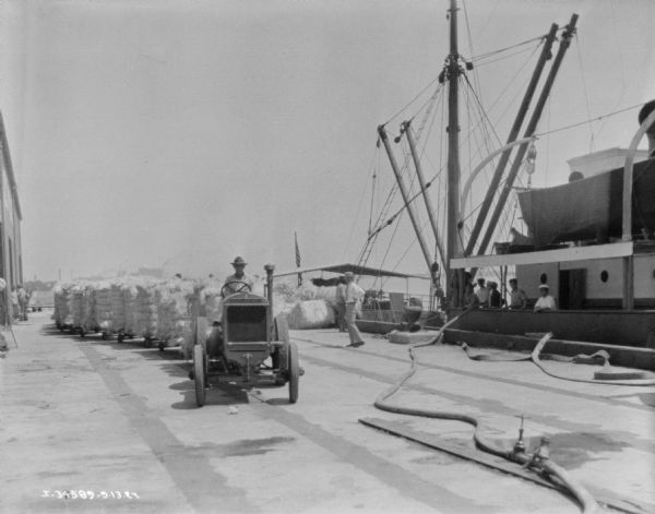 Man driving industrial tractor pulling a long set of carts stacked with bales. On the right, men are standing on a large ship at dock.
